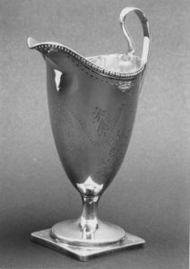 Silver creamer with a stem, handle, and delicate spout. Engraved with circular patterns and leaves.