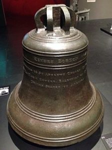 Large cast metal couch bell, inscribed with Paul Revere's name. It sits on the floor of a museum