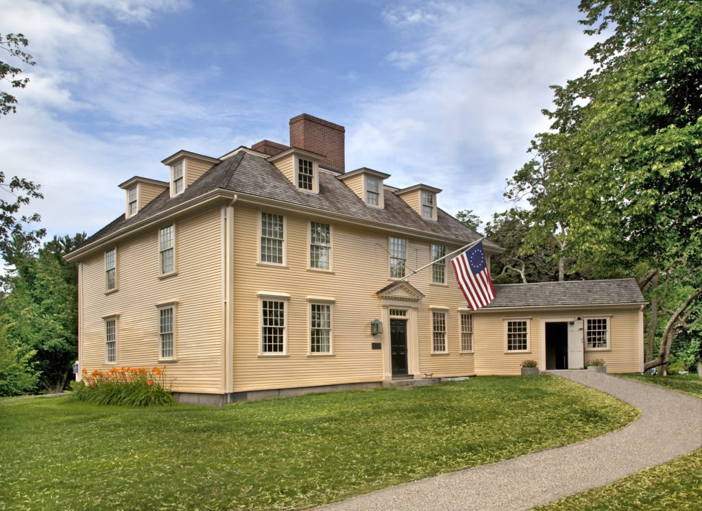 Modern photograph of a wooden building painted tan. There are gabled windows and a 13-star American Flag is displayed over the font door.