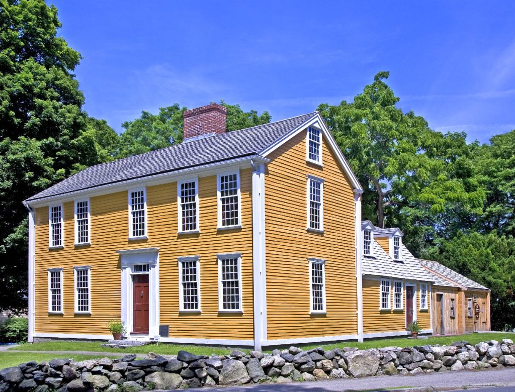 Modern photograph of a yellow painted, wooden house. The doors are painted a reddish brown color and trees line the background.