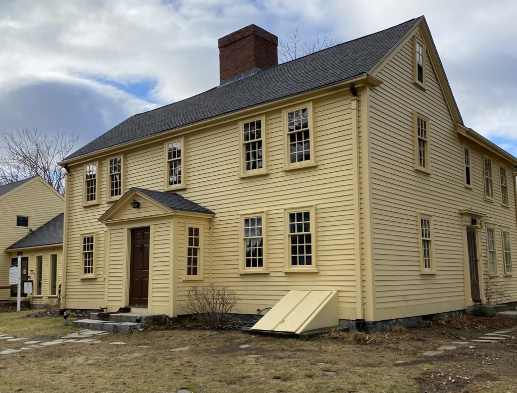 A modern photograph of a wooden house painted yellow. There is a portico around the front door and a cellar entrance visible on the right side of the image.