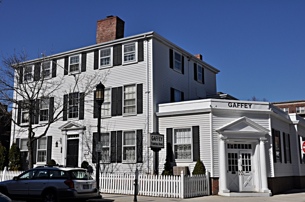 Modern photograph of a wooden house painted white with a blue sky background. The house has black shutters and reads "Gaffey," the name of the funeral home that occupies the space.