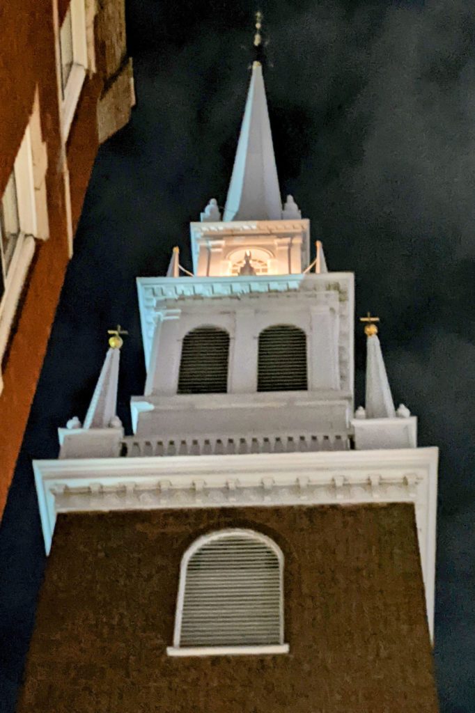 Modern photograph of a church steeple at night. The church is red brick and the steeple is wood painted white. Two lights are lit in the top of the steeple.