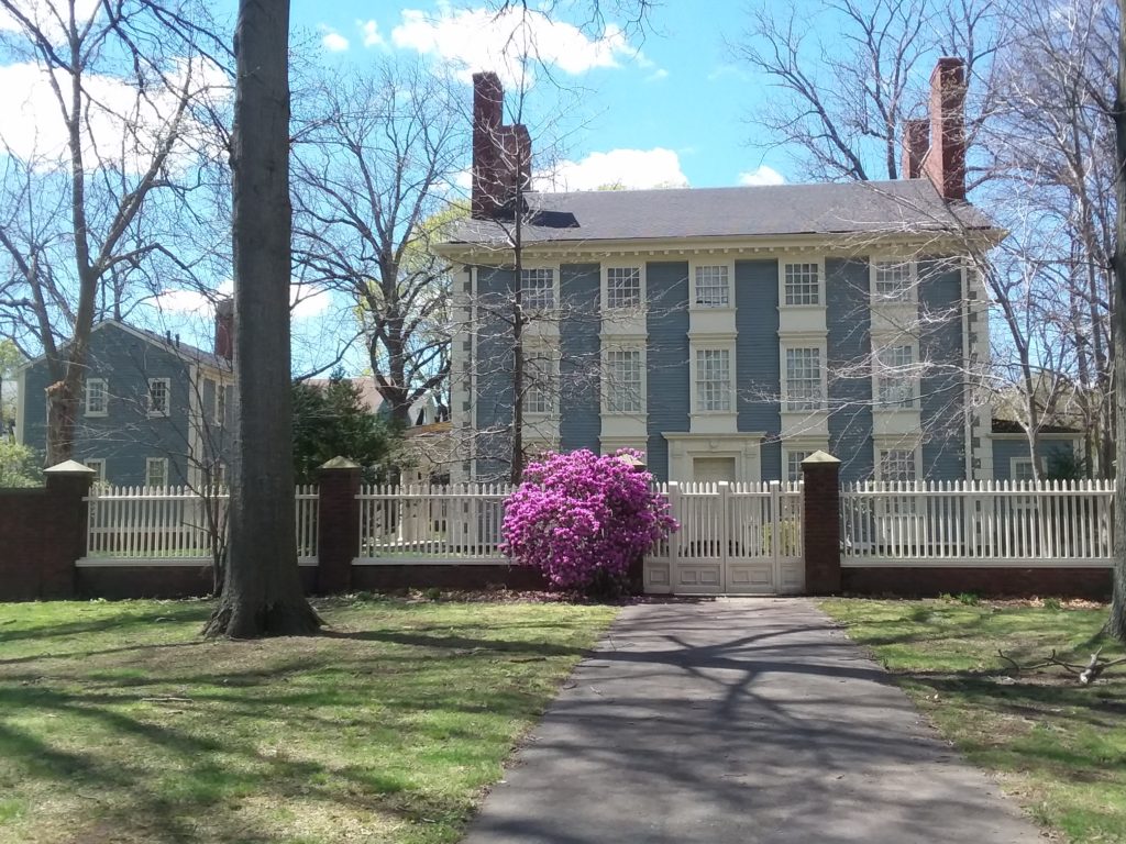 Modern photograph of a painted blue house with 14 windows. There is a white picket fence in front of the house with a paved driveway leading to the front door.