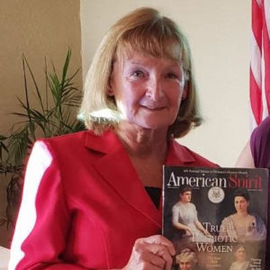 A woman with short blonde hair in a red blazer holds up a copy of "American Spirit" magazine.
