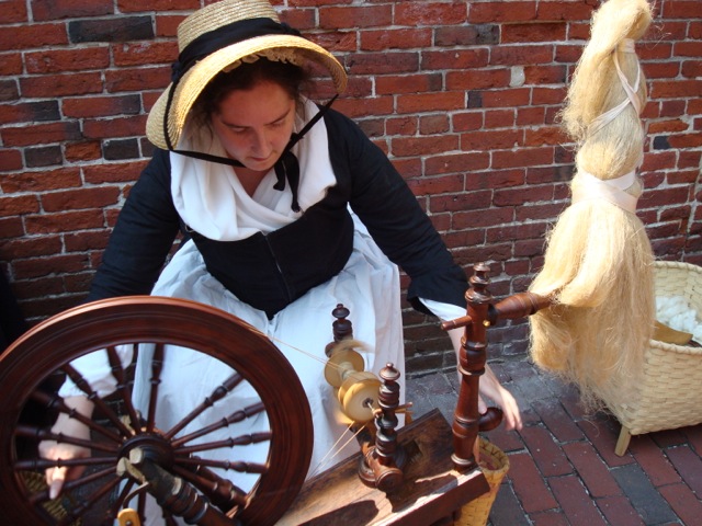 woman in colonial clothing spinning yarn on a wheel