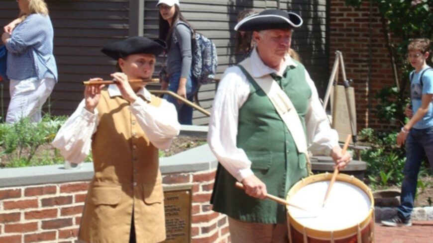 a person playing a drum next to a person playing a fife, both in colonial dress