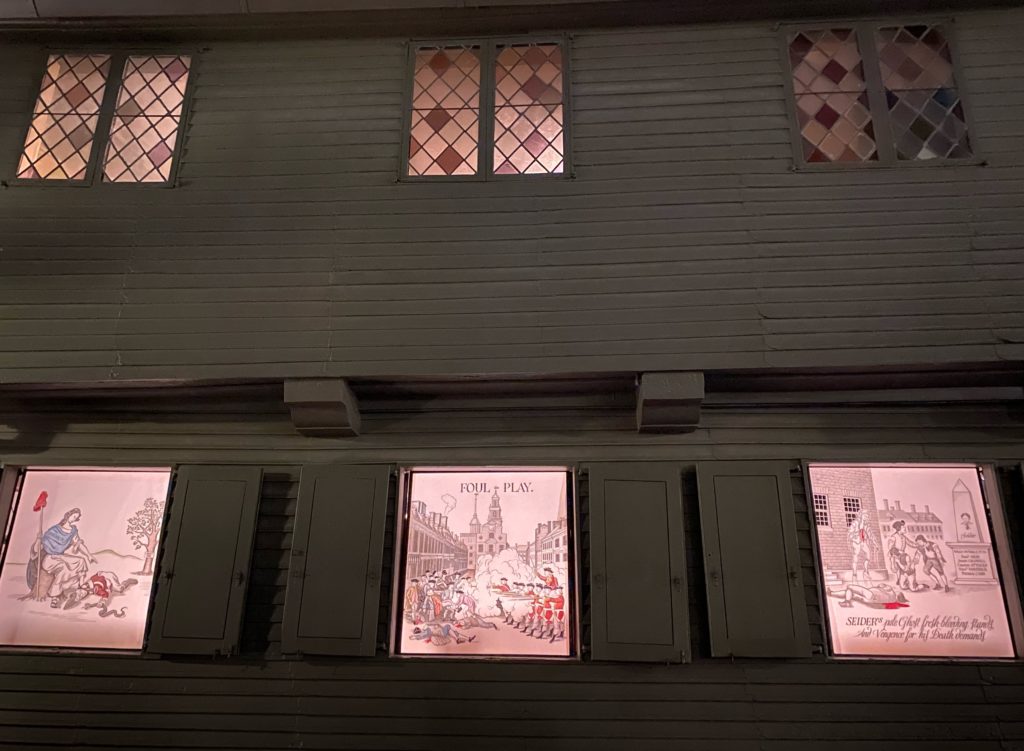 Image of the front of the Revere House, a building with dark grayish clapboards, at night. The windows are lit, and the three on the first floor show images of pre-Revolutionary events.