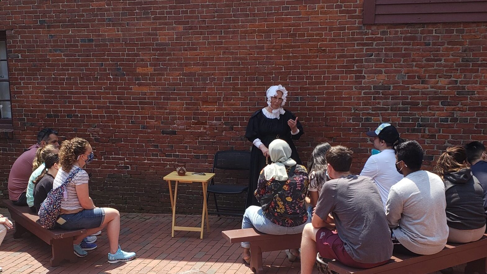 woman in colonial clothes speaking in front of people in rows on benches