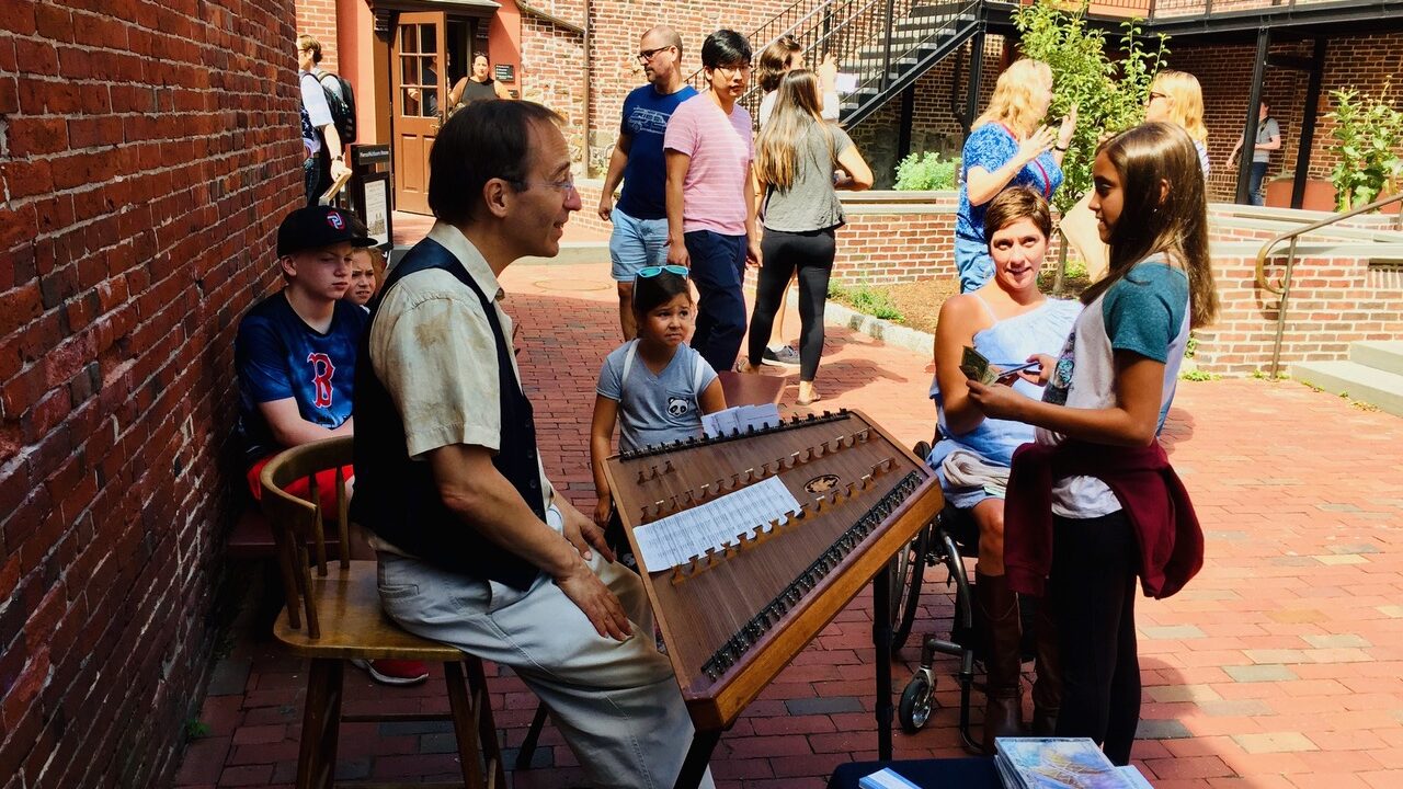 man sitting at a musical instrument called a hammered dulcimer, chatting with his audience