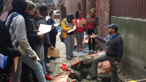 a man standing in a squareish hole a couple of feet deep, talking to several people standing outside the hole
