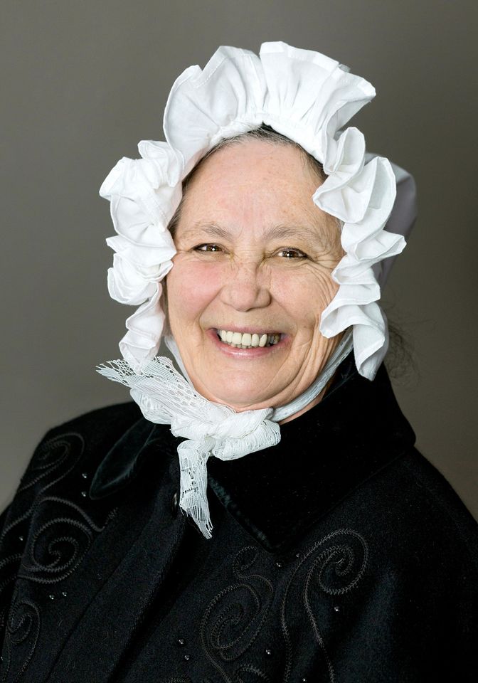 A middle-aged white woman in a white mobcap and black clothing, smiling at the camera, shown from the shoulders up.