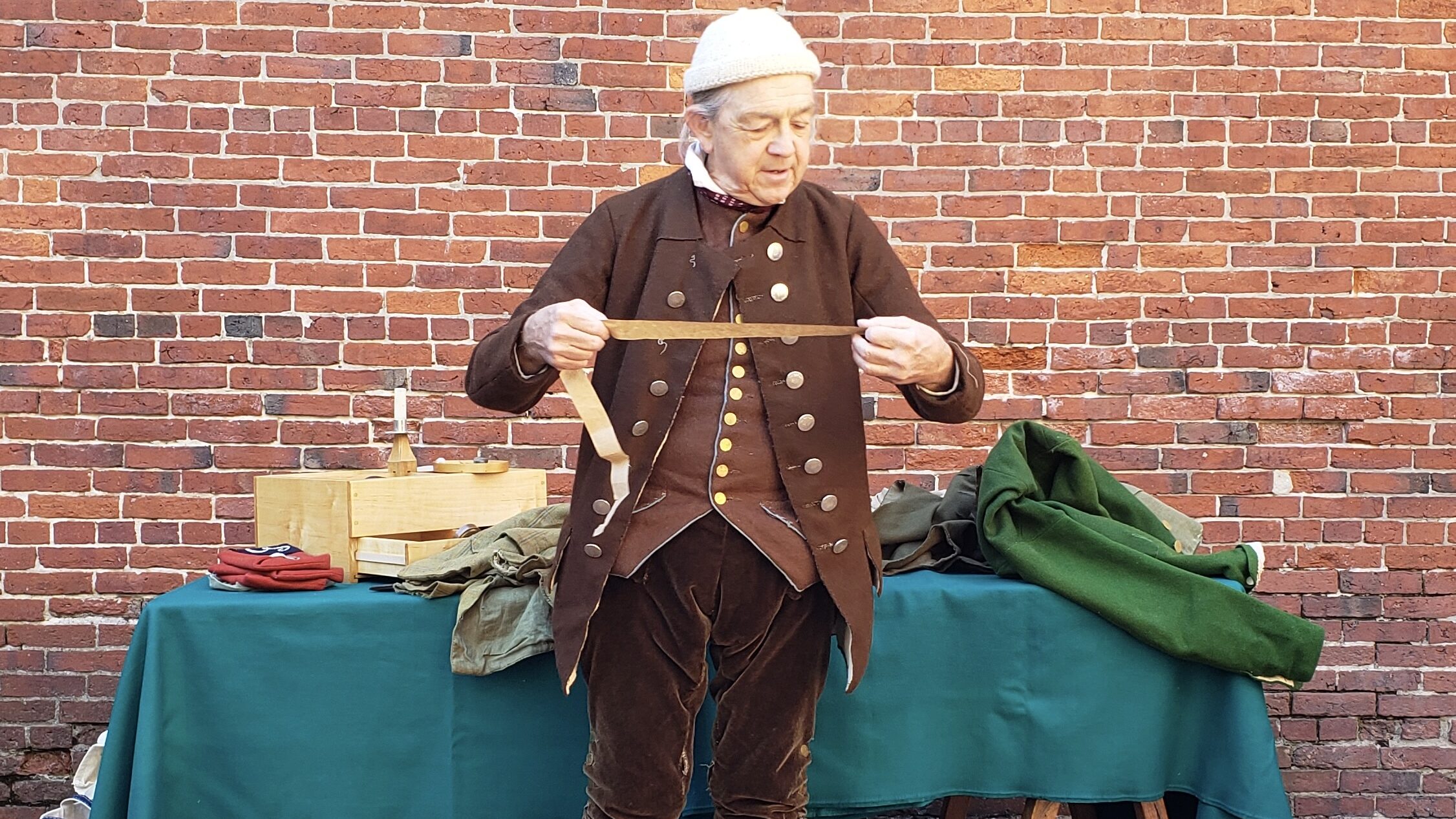 A man in brown colonial clothing holding a strip of paper and peering at it in front of a table covered in a green cloth.