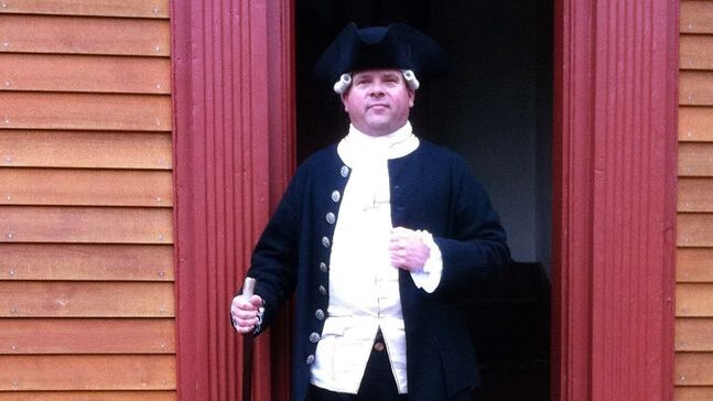 A man shown from the waist up in black and white colonial garb including a black tricorn hat. He has a walking stick in one hand, and stands in front of a doorway gazing in the general direction of the camera.
