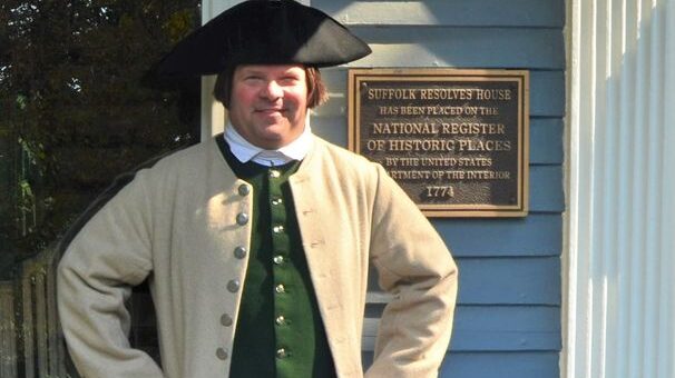 A photo of a smiling white man with earlobe length brown hair, wearing a tricorn had and 18th century clothing, with his hands on his hips. He is shown from the waist up.