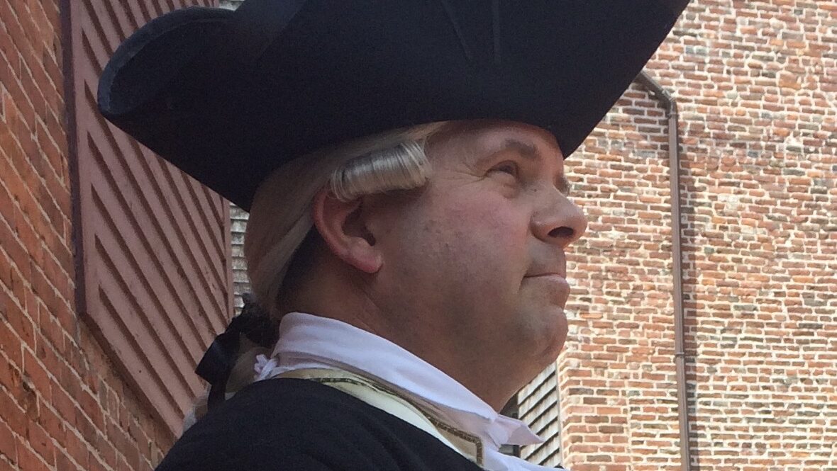 Close- up image of a man in a white wig and black tricorn hat looking determinedly ahead, in profile facing to the right.