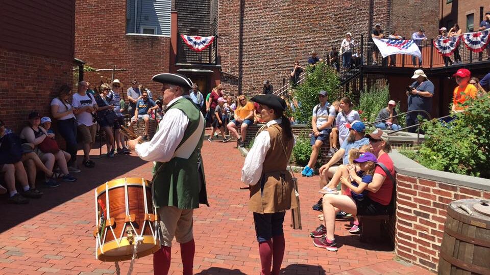Two people in 18th century clothing face to the left, away from the camera, with a large crowd of people gathered facing them. One musician has a large drum.