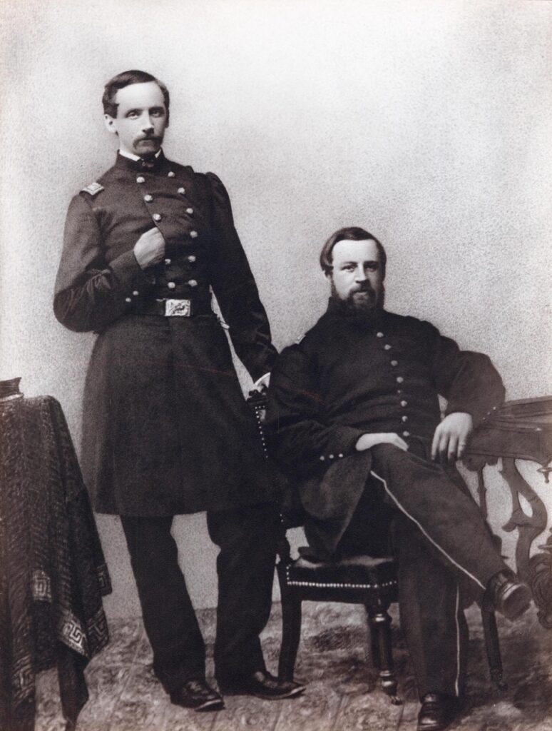 Two men wearing military uniforms, one seated and one standing next to him, in a black and white photograph.