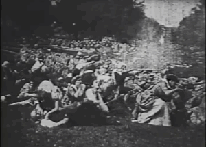 a still of a battle scene from the Midnight Ride of Paul Revere. In the scene, colonists crouch behind a stone wall, firing guns. Towards the front of the shot, a woman is holding a gun and aiming it at a british soldier offscreen