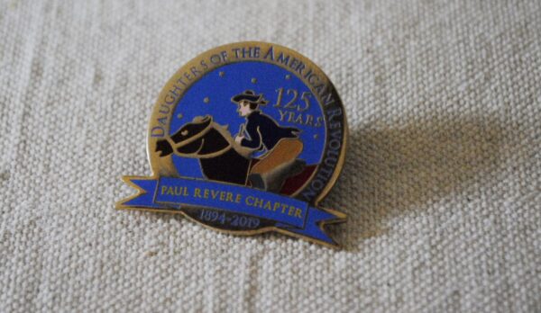 A quarter-sized metal circle pin celebrating the Daughters of the American Revolution's "Paul Revere Chapter." The center features a blue background with small stars, a picture of Paul Revere on a brown horse with a banner reading "Paul Revere Chapter", and the dates "1894–2019". The gold colored border says "Daughters of the American Revolution" in white letters, and the words "125 Years" at the top right.