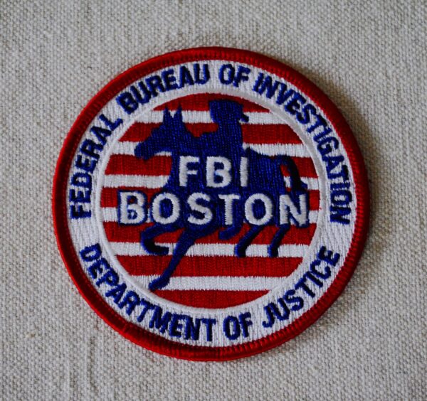 an embroidered patch with a red edge, then a white one, with blue lettering saying FEDERAL BUREAU OF INVESTIGATION at the top and DEPARTMENT OF JUSTICE on the bottom. The interior shows a silhouette of a man on a horse superimposed over red and white stripes, over which white letters say FBI BOSTON.