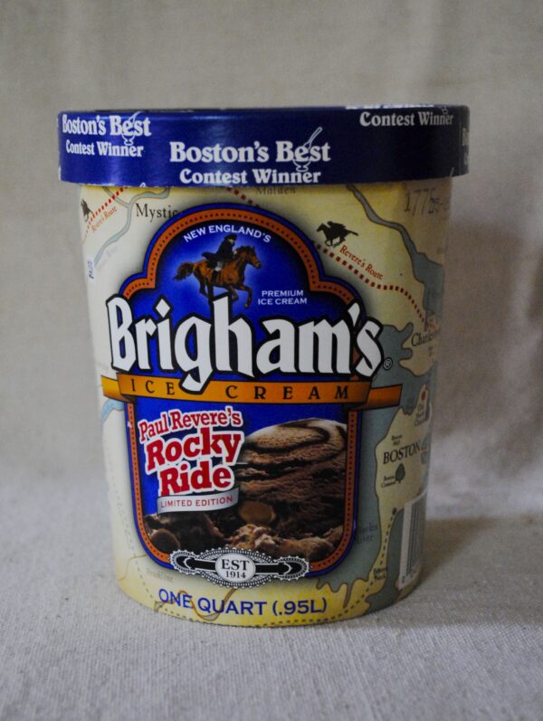 Paul Revere’s Rocky Ride is a one-quart container of Brigham Ice Cream with a pale yellow label featuring the blue Brigham logo, chocolate ice cream with chunks and caramel chips, and an illustration of Paul Revere on horseback. The background shows a map of Boston and Revere’s route to Lexington, with the Nutrition Facts panel printed on the side of the container.