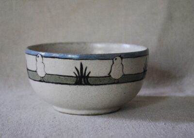 A small white ceramic bowl decorated with three horizontal stripes: blue, white, and green. Repeating stylized images of white chicks and black wheat stalks are painted onto the stripes.