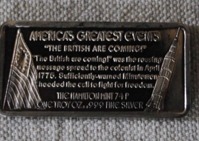 the back of a silver bar, showing an American flag and a spaceship. Text reads: "America's Greatest Events. 'THE BRITISH ARE COMING!' 'The British are coming!' was the rousing message spread to the colonist in April, 1775. Sufficiently-warned Minutemen heeded the call to fight for freedom. THE HAMILTON MINT 74P ONE TROY Ox..999 FINE SILVER"