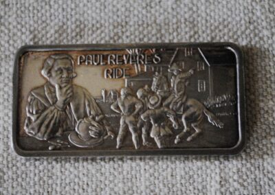 a silver bar embossed with an reproduction of the famous Copley portrait of Paul Revere and an image of Paul Revere warning a crowd on horseback. Text reads "PAUL REVERE'S RIDE."