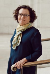 Jeannine Falino, a white woman with dark curly hair and glasses. Her hand is on a bannister and she is smiling.