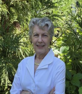 Janine Skerry, a white woman with short salt-and-pepper hair. She is smiling in front of a leafy green background.