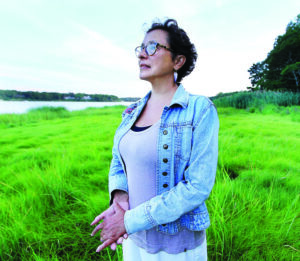 Paula Peters stands outdoors, in front of grasses and water. She has dark hair and glasses and is looking into the distance.